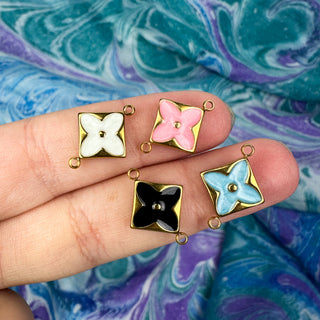 white and pink and blue and black enamel golden charms on fingers in front of a multicolor background