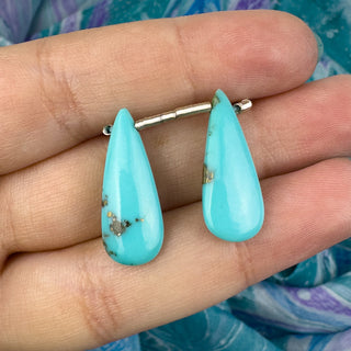 two blue turquoise teardrop earring beads on fingers with blue background