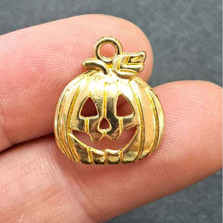 A small gold pumpkin-shaped charm with a carved jack-o'-lantern face and a loop at the top, shown resting on a person's fingers against a dark gray background.