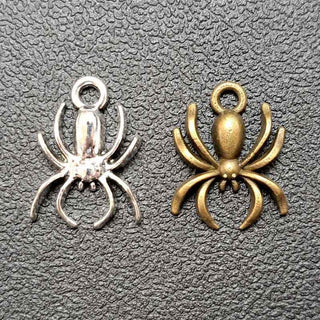 Two spider-shaped jewelry charms, one polished silver-tone and one antique bronze, displayed side-by-side on a textured dark gray background.