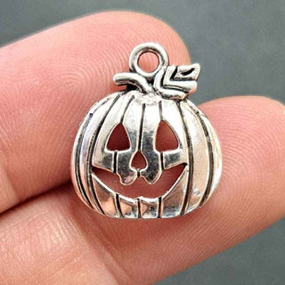 A silver pumpkin-shaped jewelry charm with a carved jack-o'-lantern face and a small loop at the top, shown resting on a person's fingers against a dark gray background.