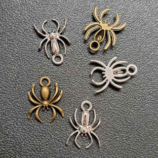 Five spider-shaped jewelry charms in both silver and antique bronze finishes, arranged on a textured dark gray surface.