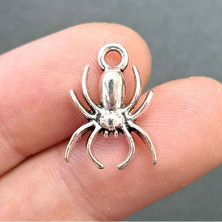 A small silver spider-shaped jewelry charm or pendant held between two human fingers against a dark gray background.