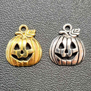 Two pumpkin-shaped jewelry charms with carved jack-o'-lantern faces, one in gold-tone metal and the other in silver-tone metal, displayed side by side on a textured gray background.